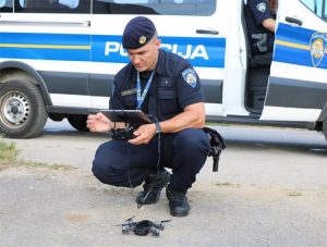 Policijski službenik u čučnju ispred policijskog vozila upravlja dronom