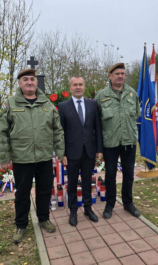 Dva muškarca u uniformama i jedan u odijelu ispred spomenika