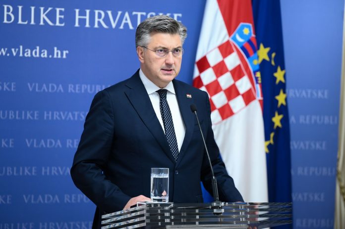 Premijer Vlade RH, Andrej Plenković. Premijer je obučen u tamnoplavo odijelo, bijelu košulju i tamnoplavu kravatu. Kraj njega stoji zastava RH