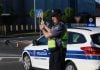 Policajac stoji i drzi znak stop, iza njega njegovo vozilo