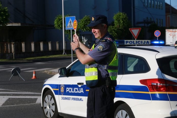 Policajac stoji i drzi znak stop, iza njega njegovo vozilo